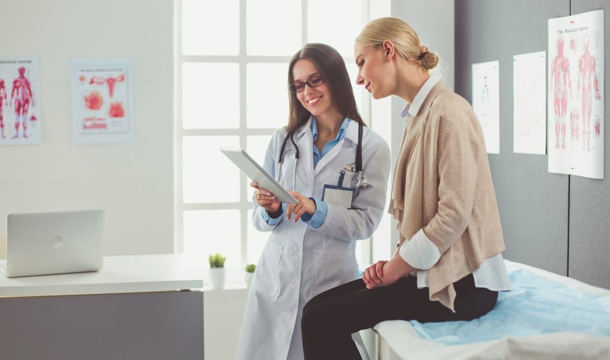 A doctor showing a patient sitting on an exam room table something on a clipboard.