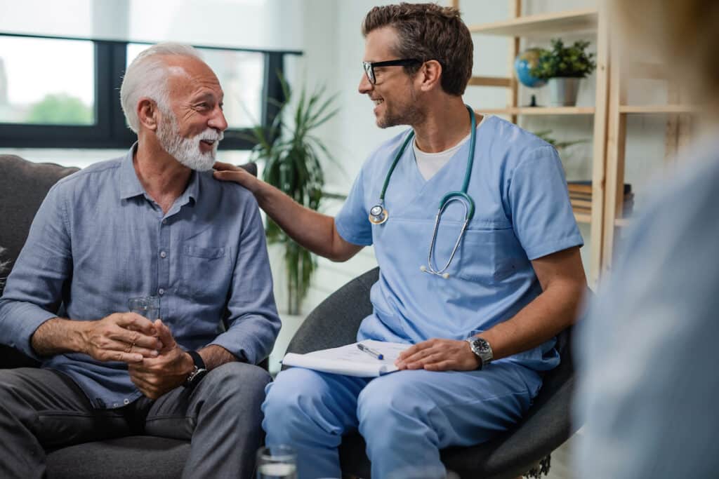 A doctor in blue scrubs places a hand on the shoulder of an elderly man as they sit and talk in an office.