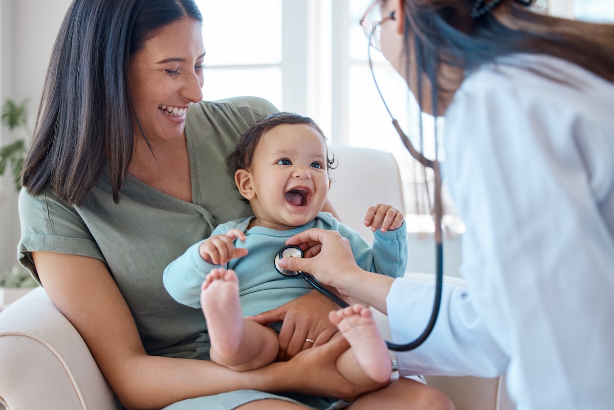 A mother holding her smiling baby as a pediatrician checks her heart with a stethoscope.