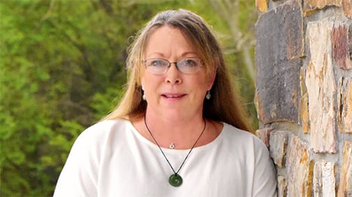 Tamara Tate's professional headshot for Healthy Connections. She wears a white shirt and green pendant and leans against a stone wall.